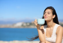 Cómo proteger tu piel del sol desde dentro Happy woman drinking coffee and smelling on the beach a sunny day