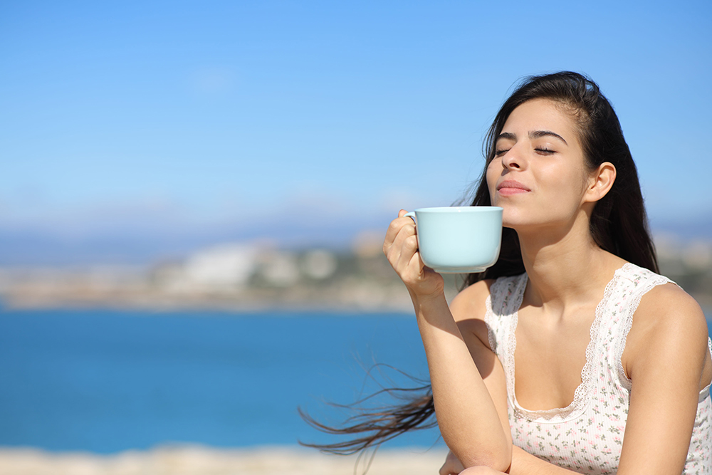 Happy woman drinking coffee and smelling on the beach a sunny day