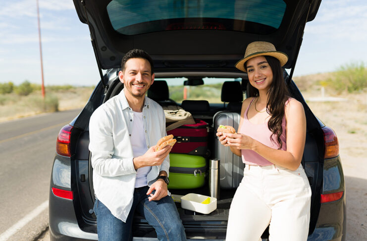 Si vas a comenzar tus vacaciones, esto te interesa Cheerful couple eating lunch in the car at the side of the road while making a long road trip together